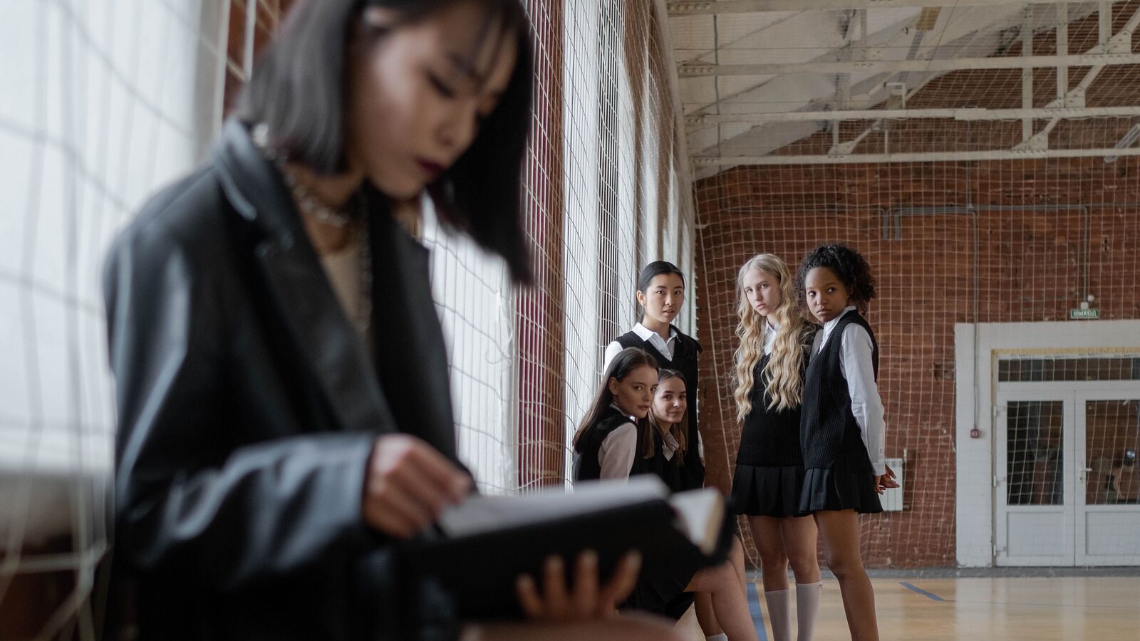 A student sitting on a bench reading from a tablet while classmates talk in the hall behind her
