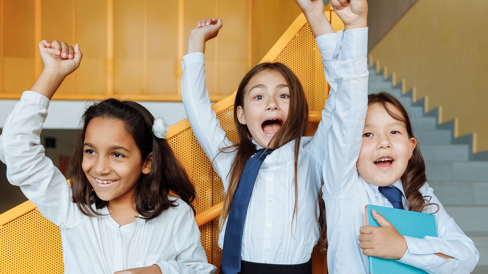 Three primary pupils cheering with their arms in the air, one holding a workbook under her arm