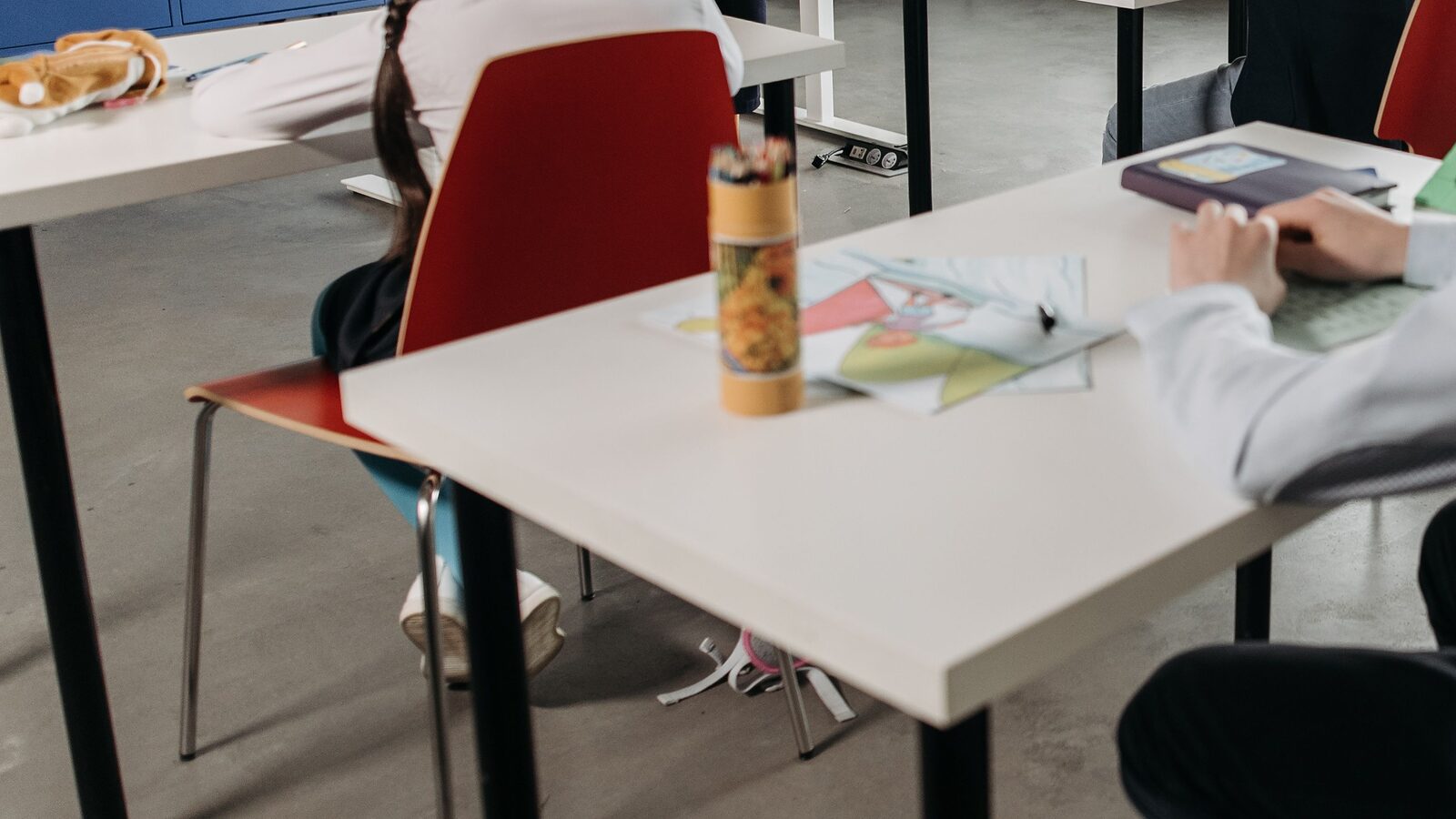 A pupil's hand resting on a workbook at a classroom desk, worksheets and a pot of pencils in front of them